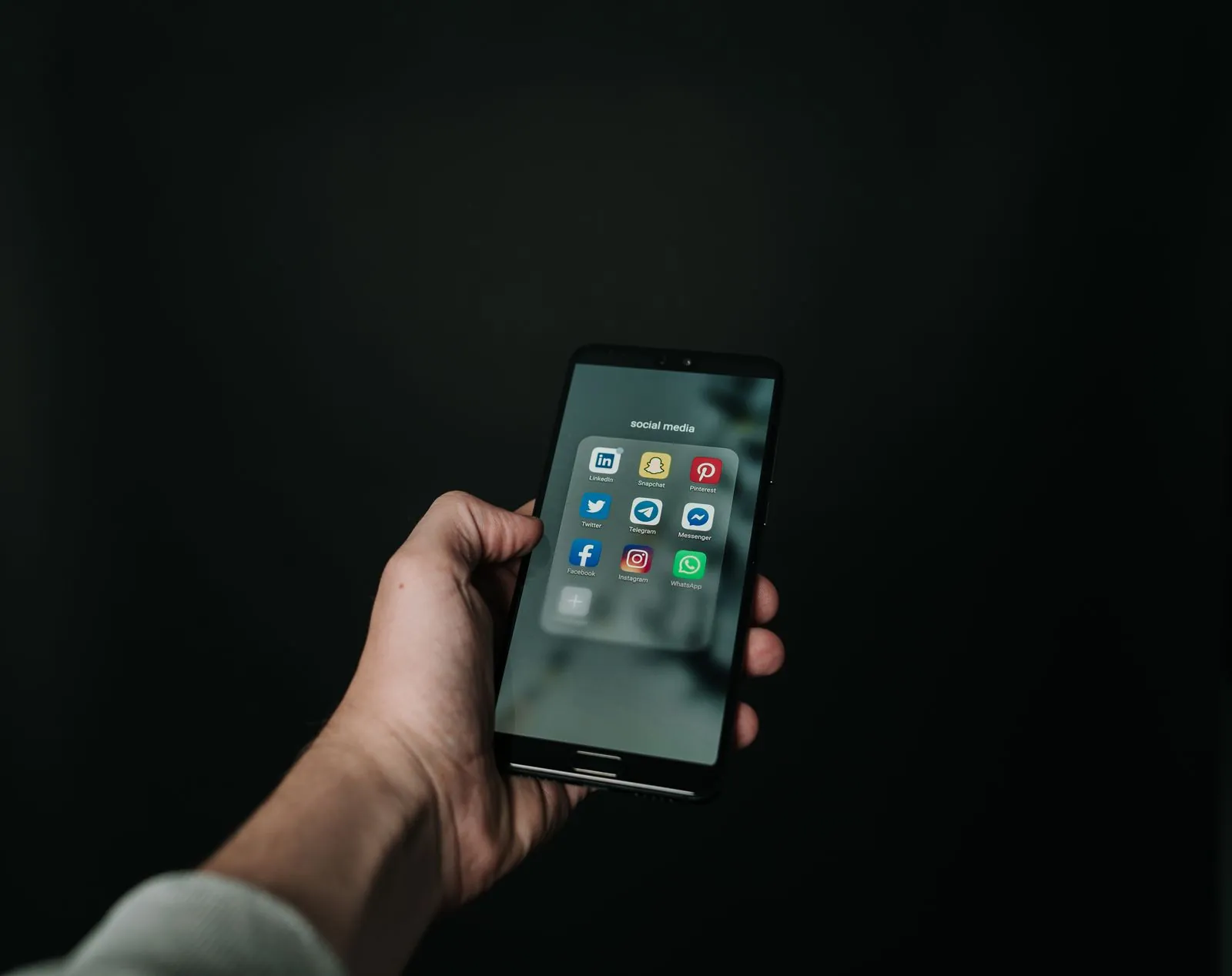 يد تمسك هاتفًا ذكيًا مفتوحًا على مجلد تطبيقات التواصل الاجتماعي، ويظهر فيه سناب شات وفيسبوك وإنستغرام وواتساب